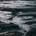 Close-up of a dark, cracked surface with shallow puddles, dimly lit and shadowed—evoking a mood of heaviness, isolation, and the absence of light or clarity.