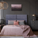 A moody, thoughtfully styled bedroom with soft pink and dark gray tones, evoking both warmth and melancholy. A neatly made bed with layered pillows and a dusty rose throw sits beneath a purple and gold abstract painting. The space is elegant but somber, visually echoing themes of early adulthood, lost innocence, and the blurred lines between comfort and control.