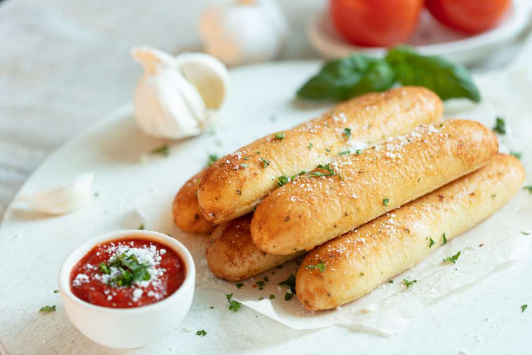 Freshly baked garlic breadsticks sprinkled with parsley and Parmesan, served with a side of marinara sauce on a white plate, with garlic cloves and tomatoes in the background.