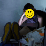 erson slumped at a cluttered desk with feet up, laptop on their lap, and a hand on their head—face covered by a yellow smiley face graphic, contrasting with the visible exhaustion and chaos around them.