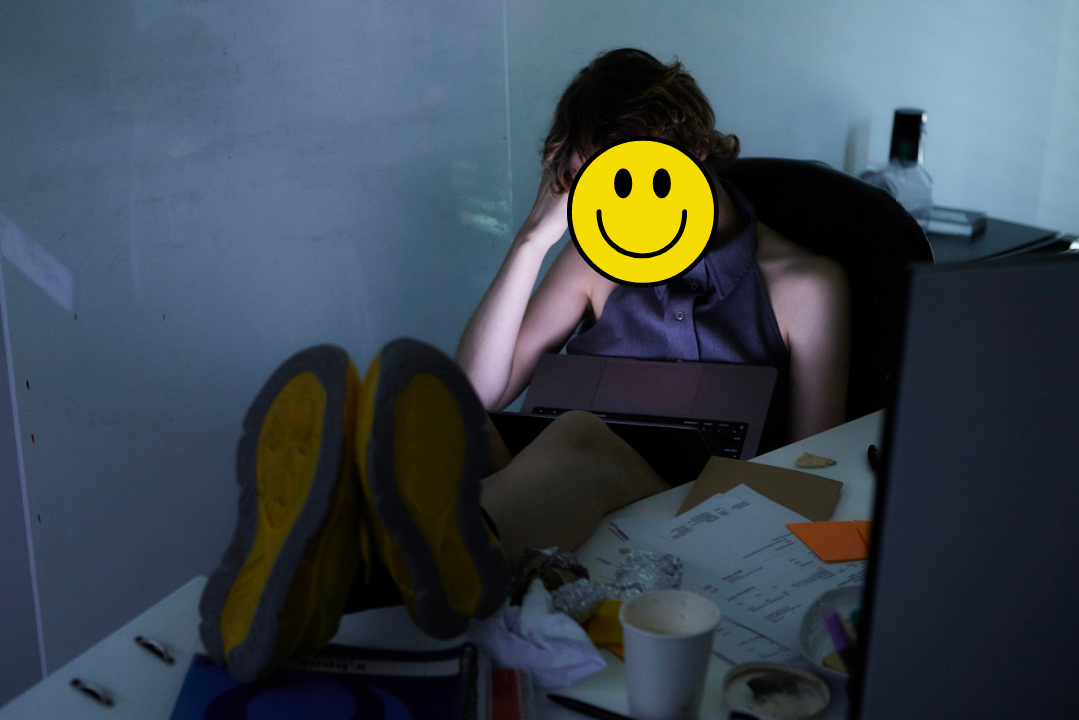 erson slumped at a cluttered desk with feet up, laptop on their lap, and a hand on their head—face covered by a yellow smiley face graphic, contrasting with the visible exhaustion and chaos around them.