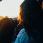 Teenage girl sitting in a car with her knees up, looking out at the sunset through the window, bathed in warm evening light.