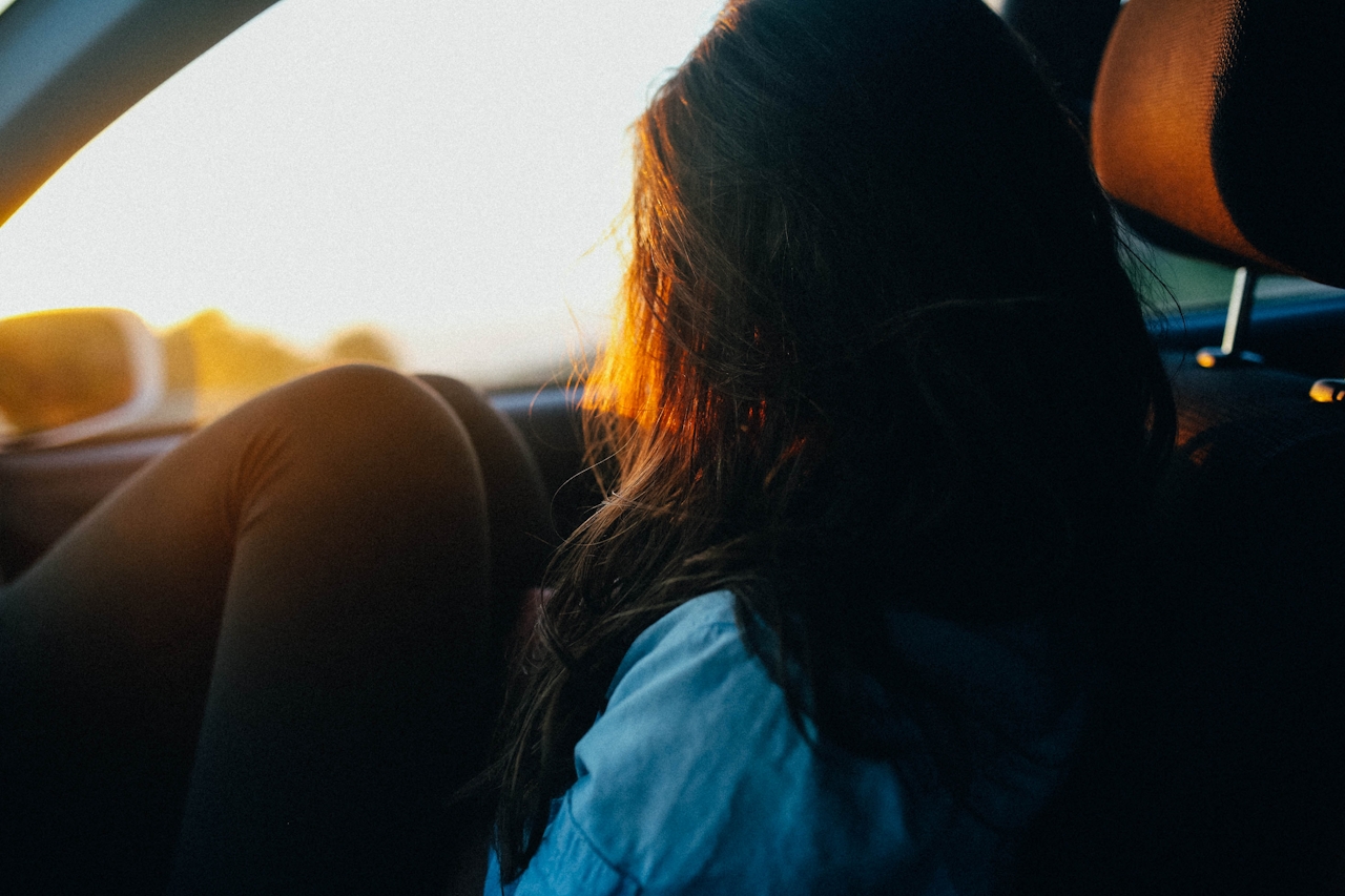 Teenage girl sitting in a car with her knees up, looking out at the sunset through the window, bathed in warm evening light.