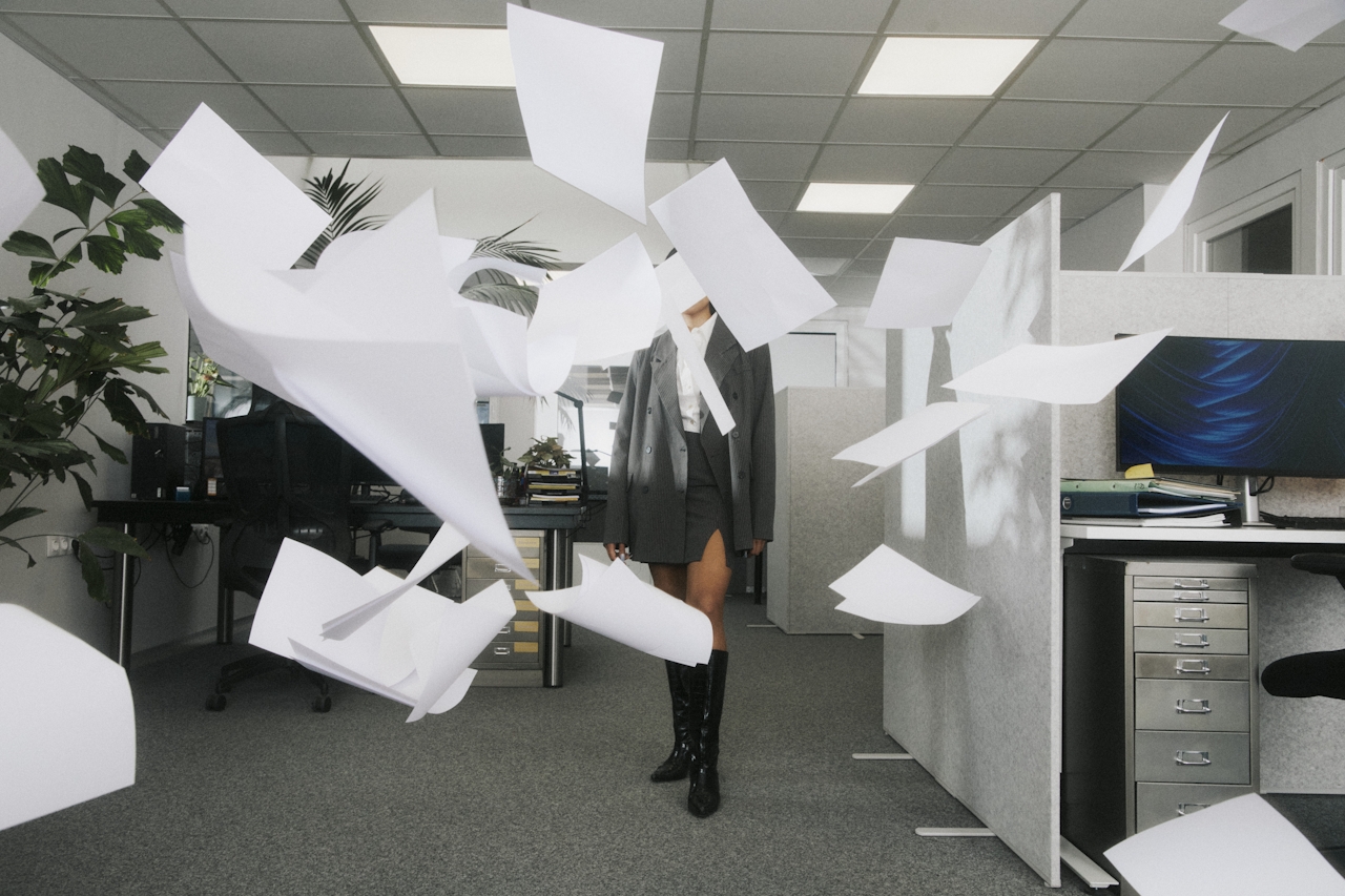 A woman in a professional outfit stands in a corporate office as sheets of paper dramatically fly through the air around her, symbolizing chaos, burnout, or a breaking point in a polished career setting.