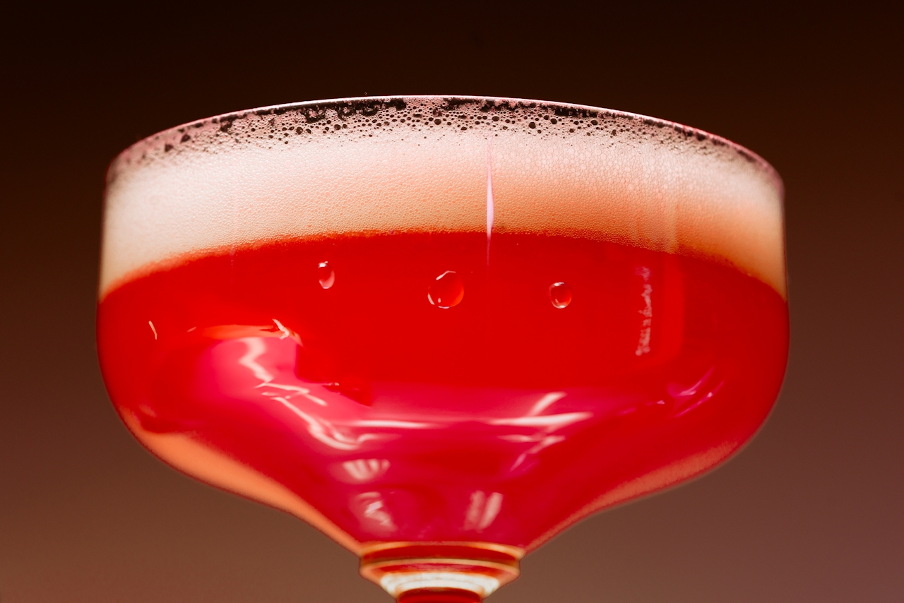 Close-up of a red sparkling cocktail in a coupe glass with a foamy head, symbolizing alcohol or celebratory drinking.