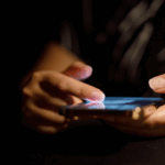 Close-up of a person using a smartphone in a dark room, highlighting the anxiety-driven habit of constantly refreshing and scrolling through feeds.