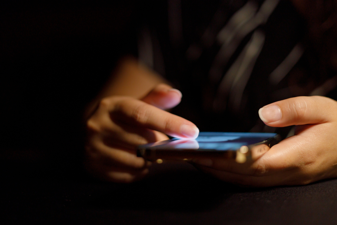 Close-up of a person using a smartphone in a dark room, highlighting the anxiety-driven habit of constantly refreshing and scrolling through feeds.