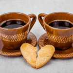 Two brown ceramic coffee cups filled with black coffee sit side by side on matching saucers, with a heart-shaped cookie broken in half placed between them—symbolizing warmth, loss, and memory.