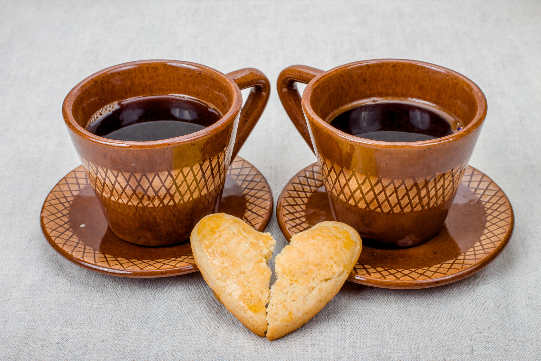 Two brown ceramic coffee cups filled with black coffee sit side by side on matching saucers, with a heart-shaped cookie broken in half placed between them—symbolizing warmth, loss, and memory.