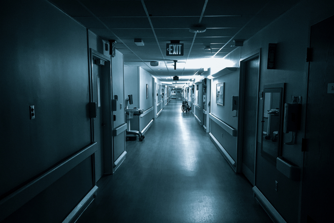 Dimly lit hospital hallway with closed doors on both sides, an empty wheelchair in the distance, and an illuminated EXIT sign above. The scene is quiet, sterile, and slightly eerie, evoking a sense of isolation or waiting.