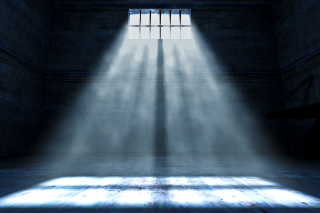 beam of light shines through high barred windows into a dark, empty cell, casting long shadows on the concrete floor—evoking the isolation and quiet heaviness of both prisons and hospital rooms.