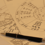 A close-up photo of printed astrology birth charts with planetary symbols and house divisions, partially overlapped, with a black pen resting on top—suggesting a reading or personal interpretation session.